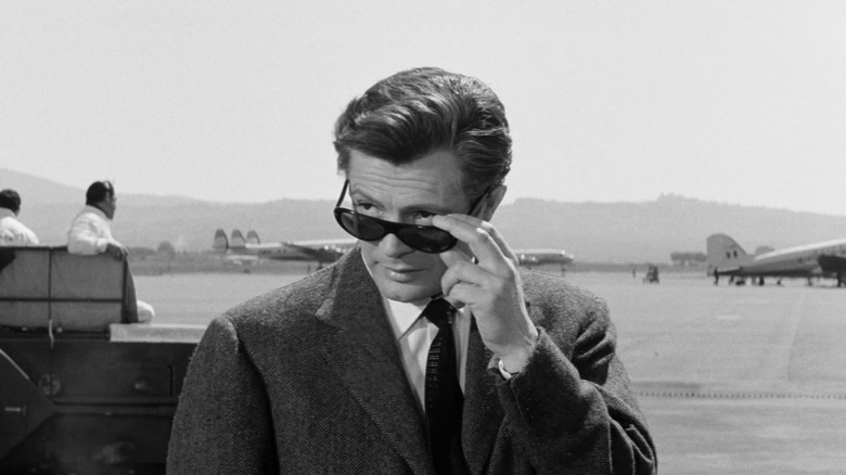 Marcello Mastroianni as Marcello Rubini in La Dolce Vita, standing against an air field in his suit and sunglasses, looking over his sunglasses slightly.
