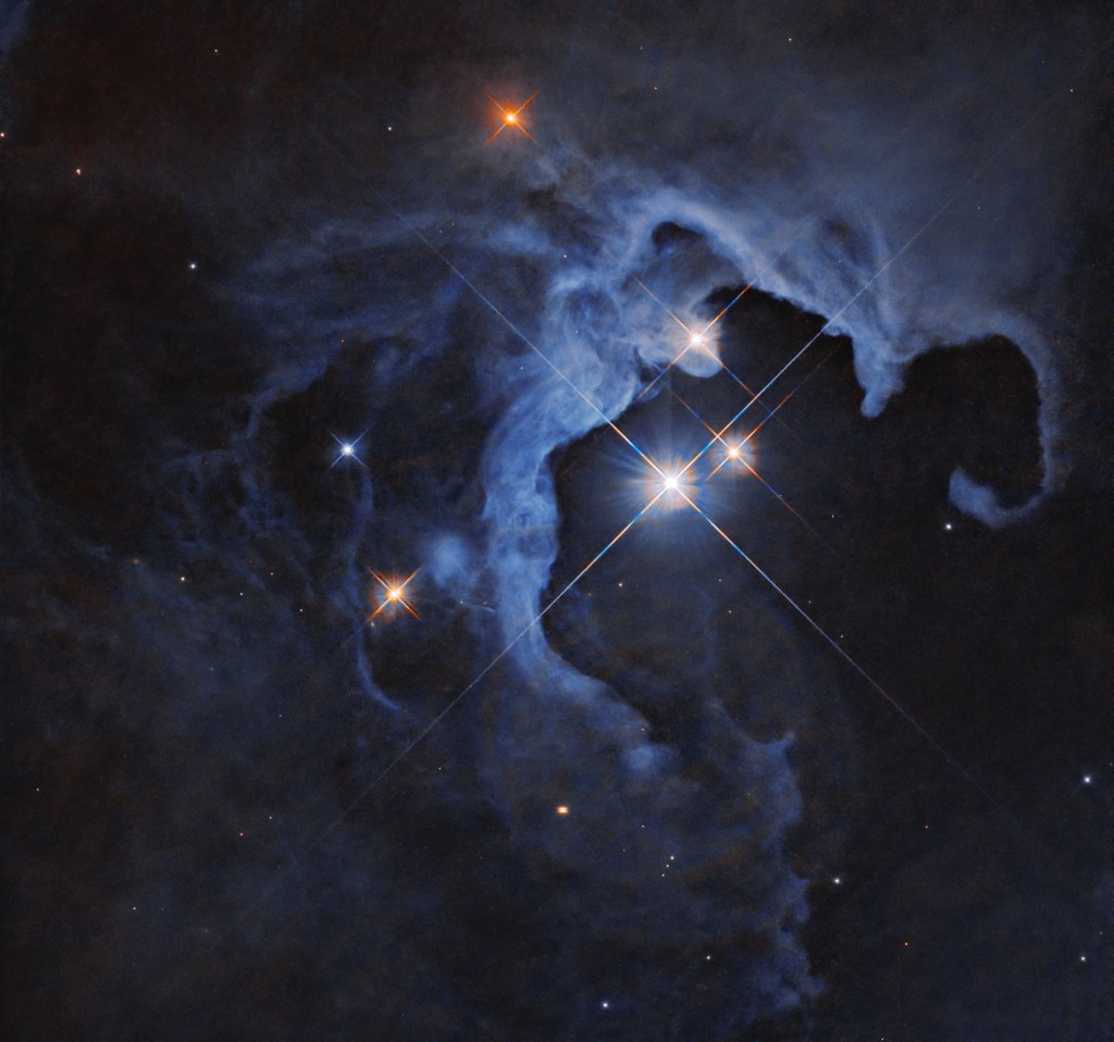 A glittering cosmic geode, a trio of dazzling stars blaze from the hollowed-out cavity of a reflection nebula.