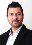 A man with short dark hair and a trimmed beard is wearing a dark blazer over a light checkered shirt, standing in front of a plain white background.