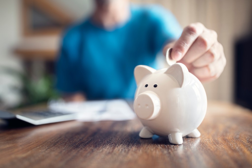 A person's hand inserting a coin into a white piggy bank on a wooden table.