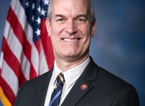 A head and shoulders portrait of a bald white man in a blue black and gold tie. He is smiling and standing in front of an American flag.