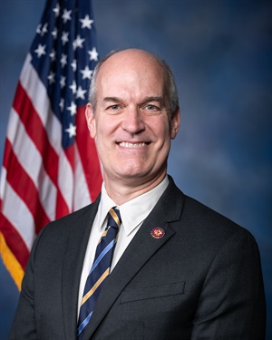 A head and shoulders portrait of a bald white man in a blue black and gold tie. He is smiling and standing in front of an American flag.