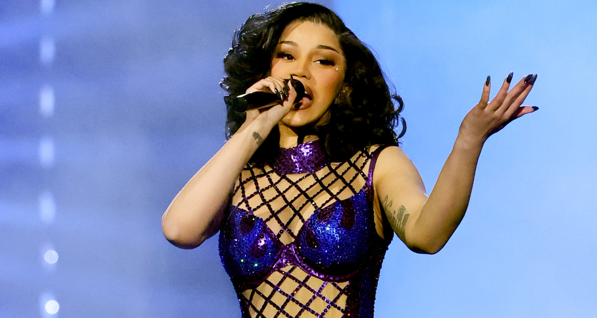 Cardi B performs on stage in a sparkling purple outfit, holding a microphone, with expressive hand gestures.