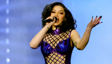 Cardi B performs on stage in a sparkling purple outfit, holding a microphone, with expressive hand gestures.