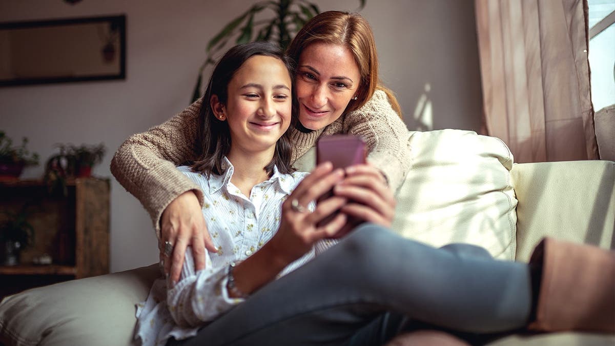 Mother and daughter sit on couch looking at smartphone together