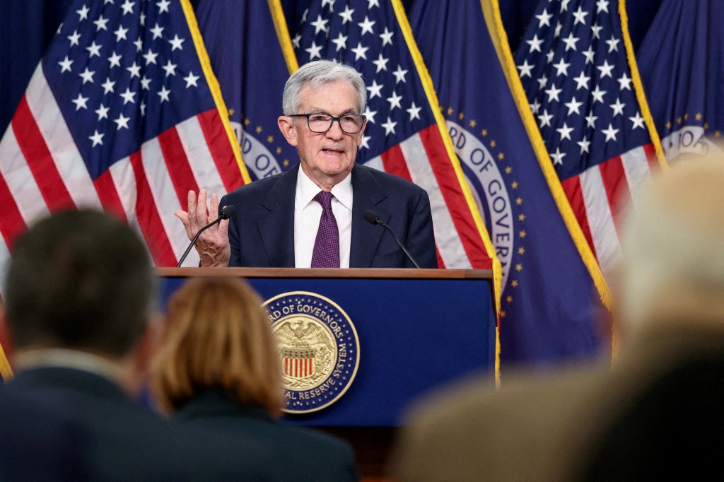 Jerome Powell speaking at a press conference with US flags in the background.