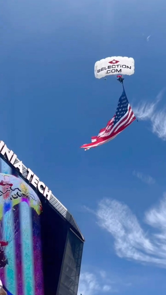 Skydiver carrying an American flag and a parachute with "SELECTION.COM" written on it, descends towards the Virginia Tech scoreboard.