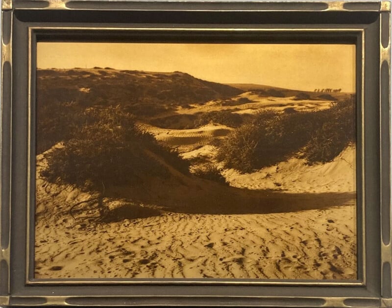 A sepia-toned photograph in a dark frame shows sand dunes with sparse vegetation under a clear sky, casting long shadows across the undulating landscape.