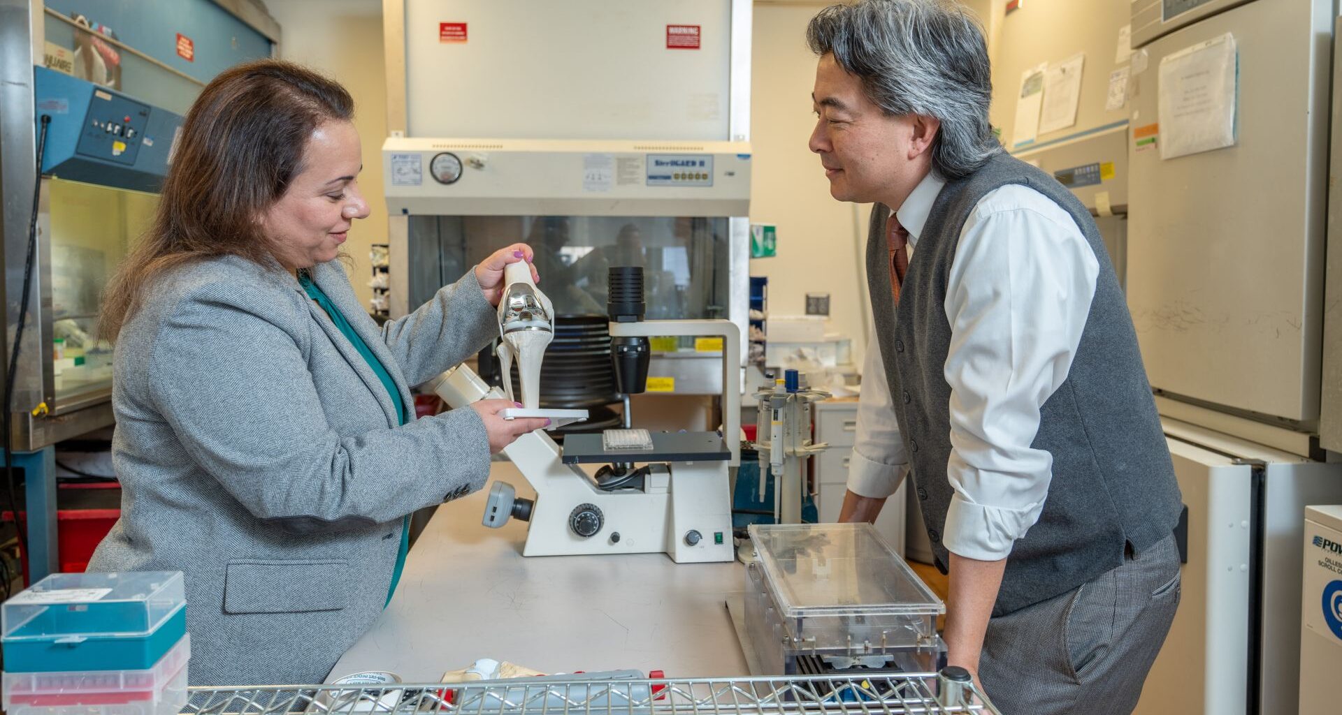 A man with long gray hair wearing a brown vest and white shirt speaks with a woman with long brown hair as they both look at various prosthetic limbs.