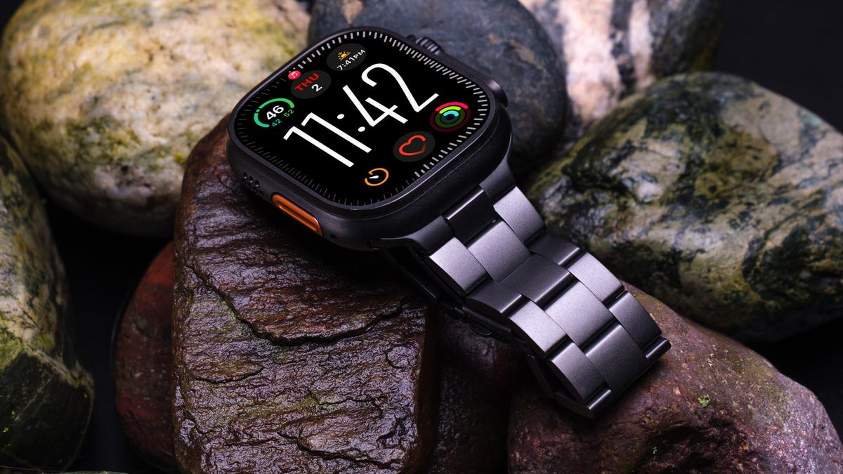 An Apple Watch Ultra 3 with a titanium link-style band lying on wet rocks.