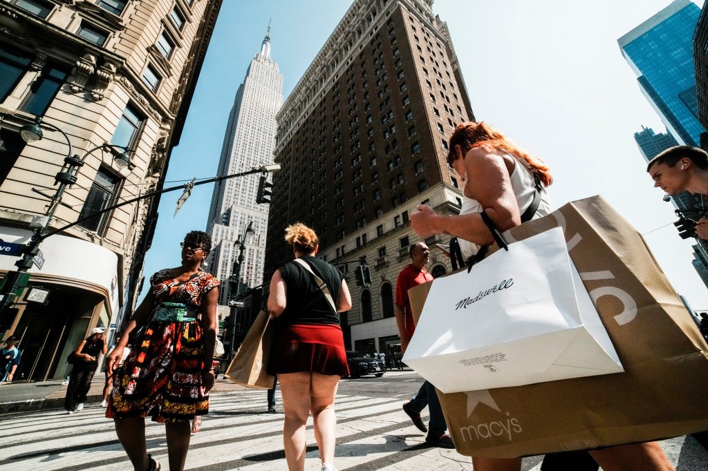 A woman carrying shopping bags walks down a street in Manhattan, with the Empire State Building visible in the background.