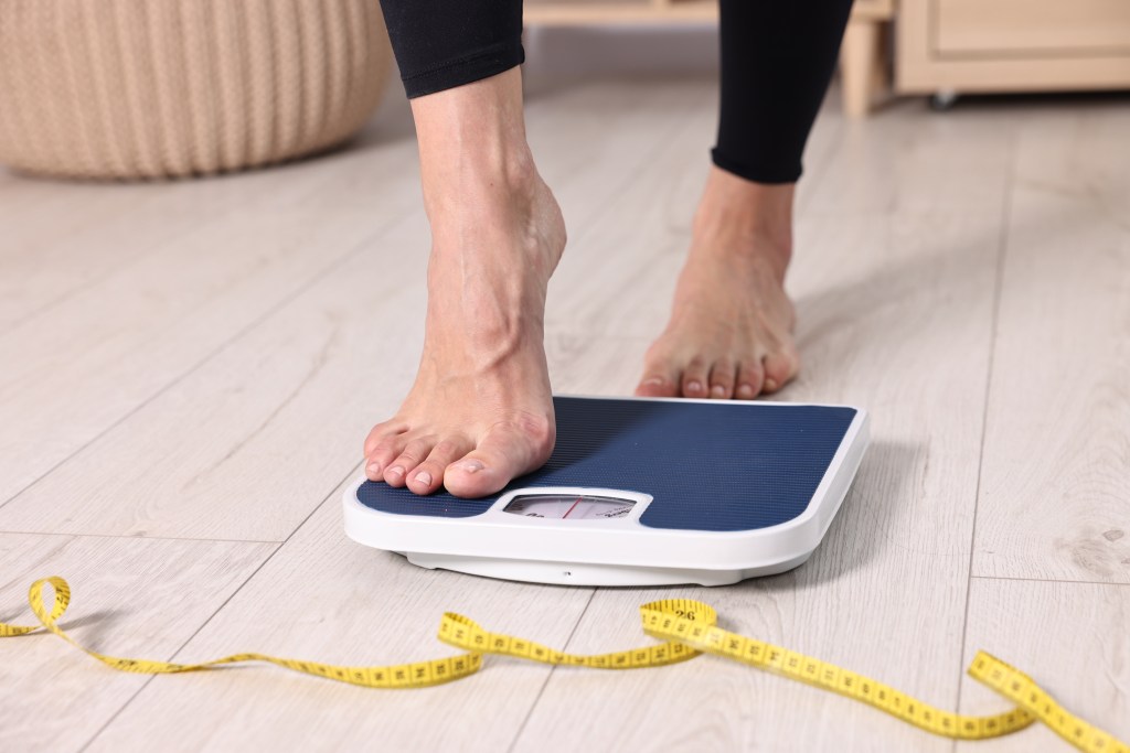 A woman stepping on a bathroom scale, with a yellow measuring tape on the floor beside it.