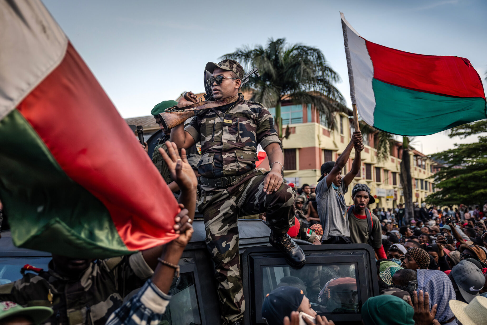 Luis Tato, “Madagascar’s Gen Z Protests,” for Agence France-Presse. Protesters cheer and wave flags outside City Hall as members of the CAPSAT military unit ride on an armored vehicle. Antananarivo, Madagascar, October 11, 2025.