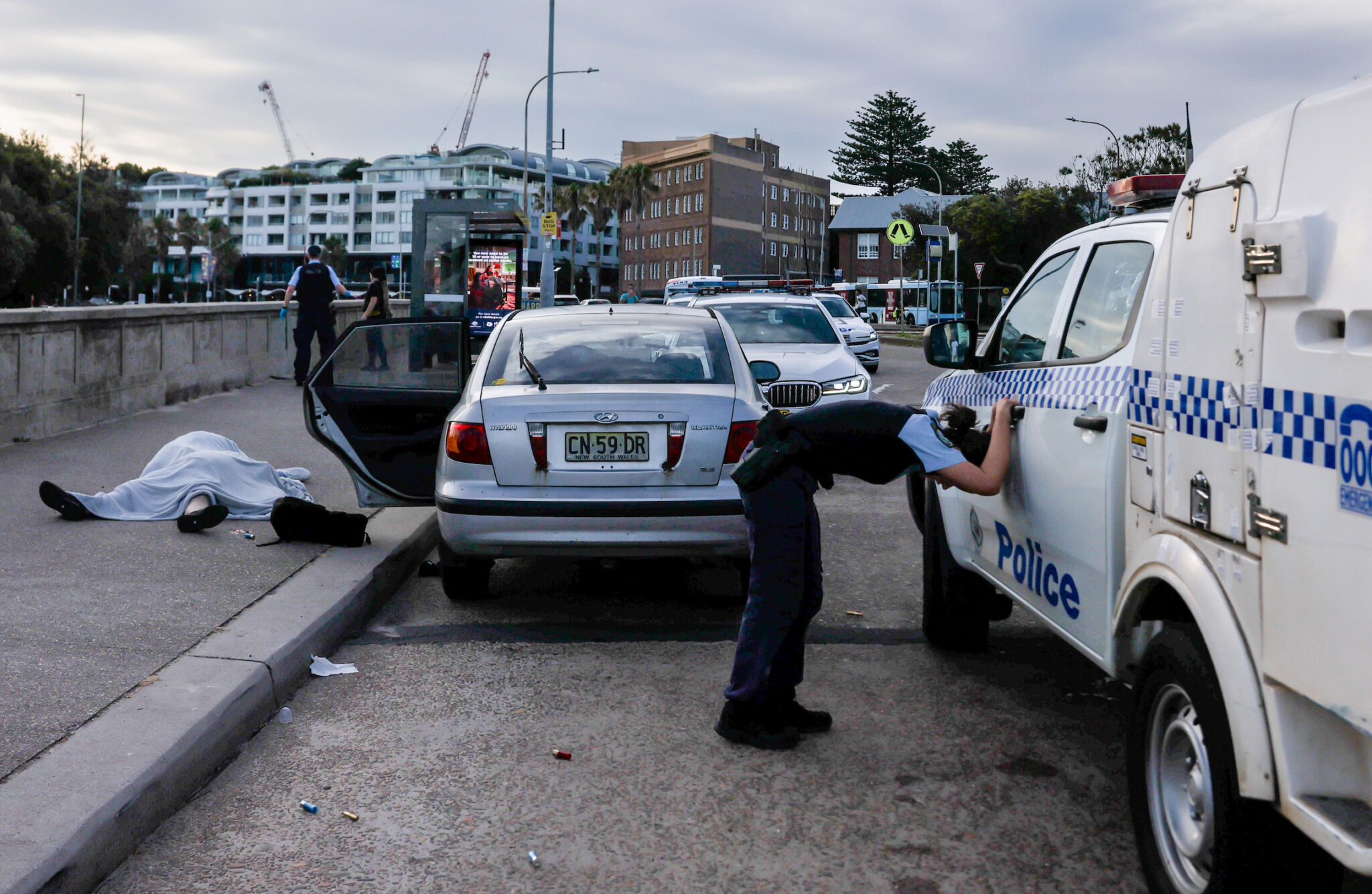 Edwina Pickles, “Bondi Beach Terror Attack,” for the Sydney Morning Herald. An overwhelmed police officer leans over near the bodies of Boris (69) and Sofia (61) Gurman. During the Bondi Beach Terror Attack, the couple were killed while attempting to disarm one of the shooters. Sydney, Australia, December 14, 2025.