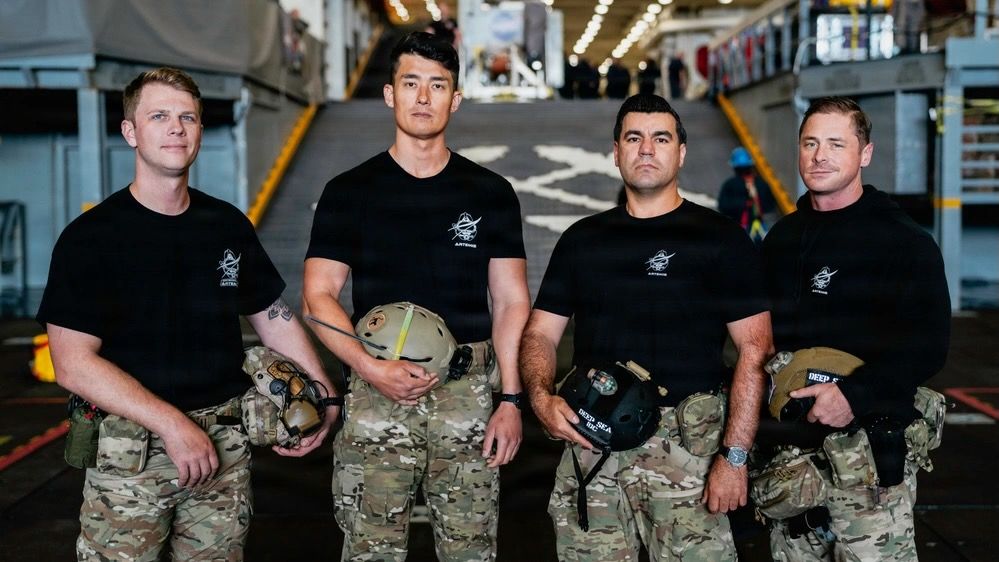 four young military men wearing black t-shirts and camouflage pants stand on the deck of a ship at sea