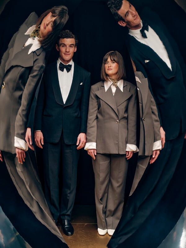 A man and woman in formal suits stand side by side, facing forward, with their distorted reflections surrounding them on both sides, creating a surreal, mirrored effect.
