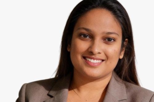 A woman with long brown hair wears a brown blazer.