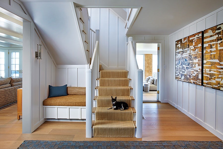 A modern staircase with beige carpet runners and white railings, featuring a small black and white dog lying on the steps. To the left of the stairs is a built-in bench with a brown cushion and a blue pillow, set against white paneled walls. On the right wall, there are two large abstract art pieces in brown, white, and black tones. The floor is light wood, and a blue patterned rug is partially visible at the bottom of the image. In the background, a doorway leads to a bright room with a sofa and striped pillows.