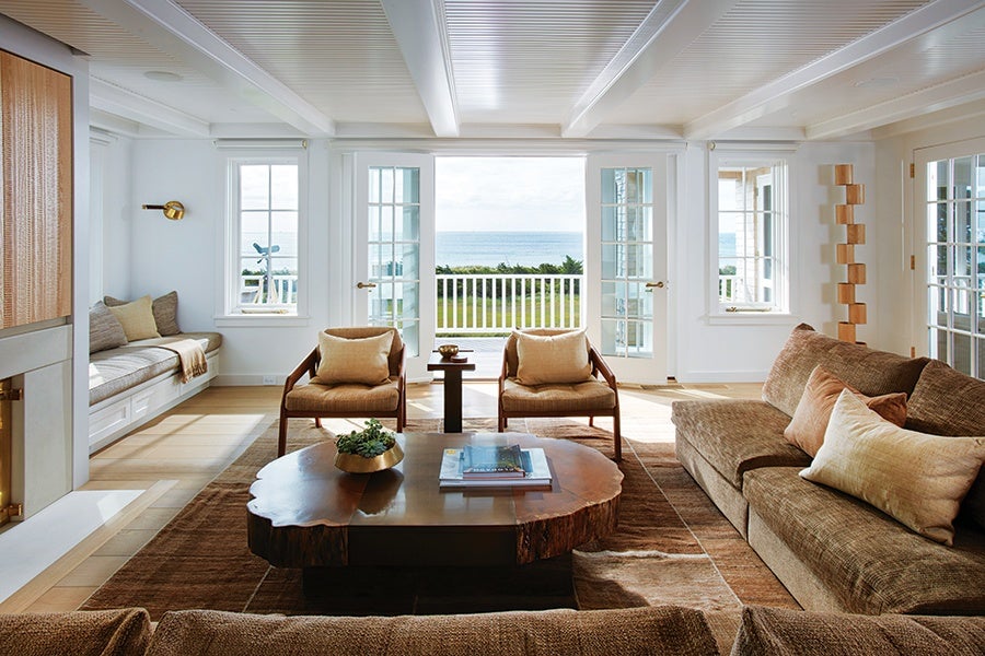 Living room with a large brown sectional sofa adorned with beige and brown pillows, two wooden armchairs with beige cushions, and a rustic wooden coffee table with books and a small plant. The room features white walls, large windows, and French doors that open to a balcony with a view of the ocean and greenery. A built-in bench with cushions is positioned under one window, and a modern wooden floor lamp stands near the sofa. The space is bright with natural light and has a cozy, coastal aesthetic.