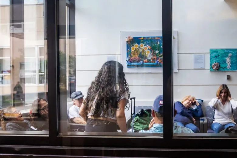 A group of people sit and wait in a room with artwork on the walls, viewed through a glass window. One woman stands while others are seated, some appearing to rest or talk.