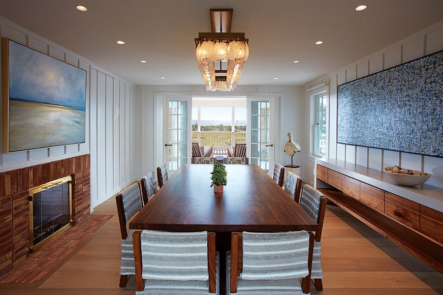 Dining room with a large rectangular wooden table surrounded by eight striped upholstered chairs. A modern rectangular chandelier with multiple glass globes hangs above the table. The room features white paneled walls, a brick fireplace with a brass frame on the left, and a long wooden sideboard with a white countertop on the right. Two large abstract paintings hang above the fireplace and sideboard. French doors at the far end open to a balcony with outdoor seating and a view of greenery and water in the distance.
