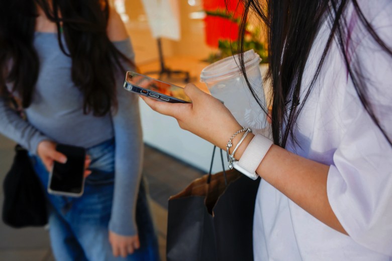 A close-up view an unidentifiable teenager holding cell phones in their hand as they hold a Starbucks drink and a shopping with the other hand. Another unidentifiable teenager also holding a cell phone is seen in the background.