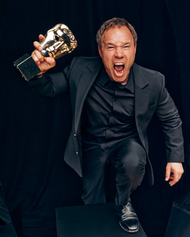 A man in a dark suit poses energetically, holding up a BAFTA trophy and shouting with excitement against a black background.