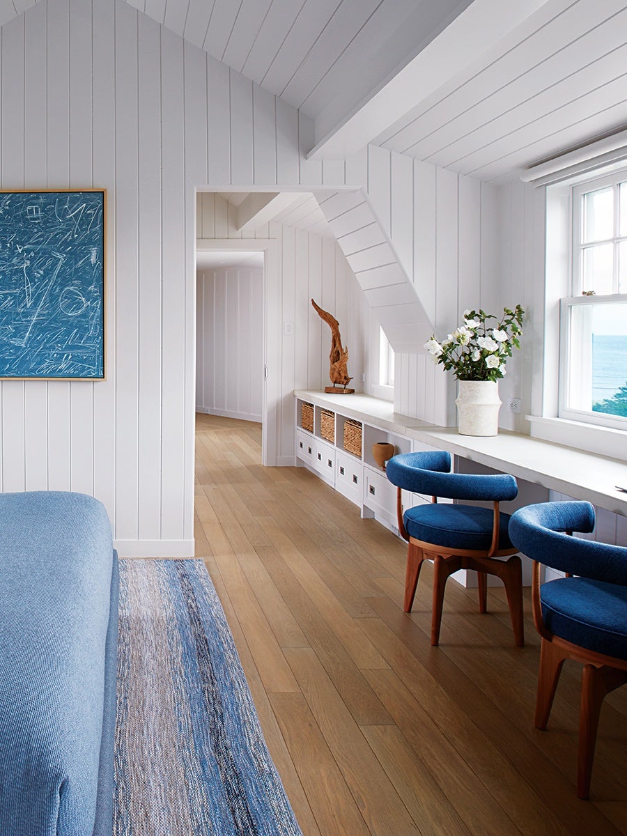 White-paneled room with light wood flooring featuring a long built-in desk under a window overlooking the ocean. Two blue upholstered chairs with wooden legs are positioned at the desk. A white vase with white flowers sits on the desk. On the left side, a blue abstract painting hangs on the wall above a blue upholstered sofa and a blue and beige striped rug. A wooden sculpture is displayed on a built-in shelf along the wall.