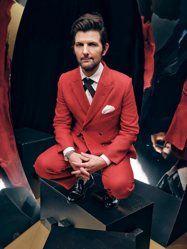 A man in a red suit with a white dress shirt and patterned tie crouches on a black geometric platform, surrounded by abstract mirrored surfaces reflecting his image.