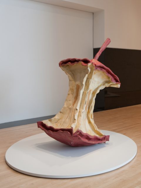 Large sculpture of a partially eaten apple core with a stem, displayed on a round white platform in a gallery setting.