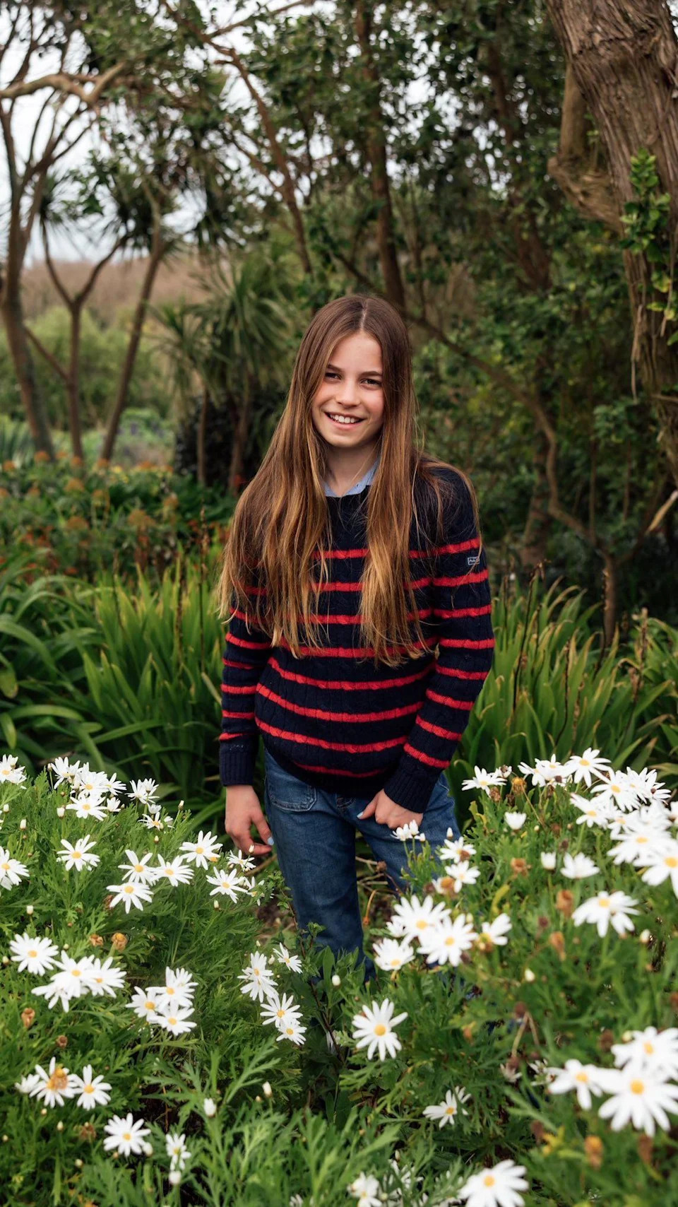 Photograph shows Charlotte wearing a black and red striped jumper and blue denim jeans, standing in a flowery field while smiling at the camera.