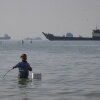 A man stands in the water, appearing to fish, as bulk carriers, cargo ships, and service vessels line the horizon in the Strait of Hormuz off Bandar Abbas, Iran, April 27.