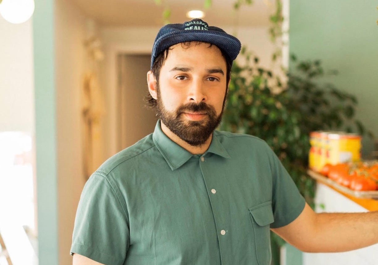 A man in a green button up short sleeve shirt and a flat capped hat stands in a restaurant with one hand on the counter in this profile photo