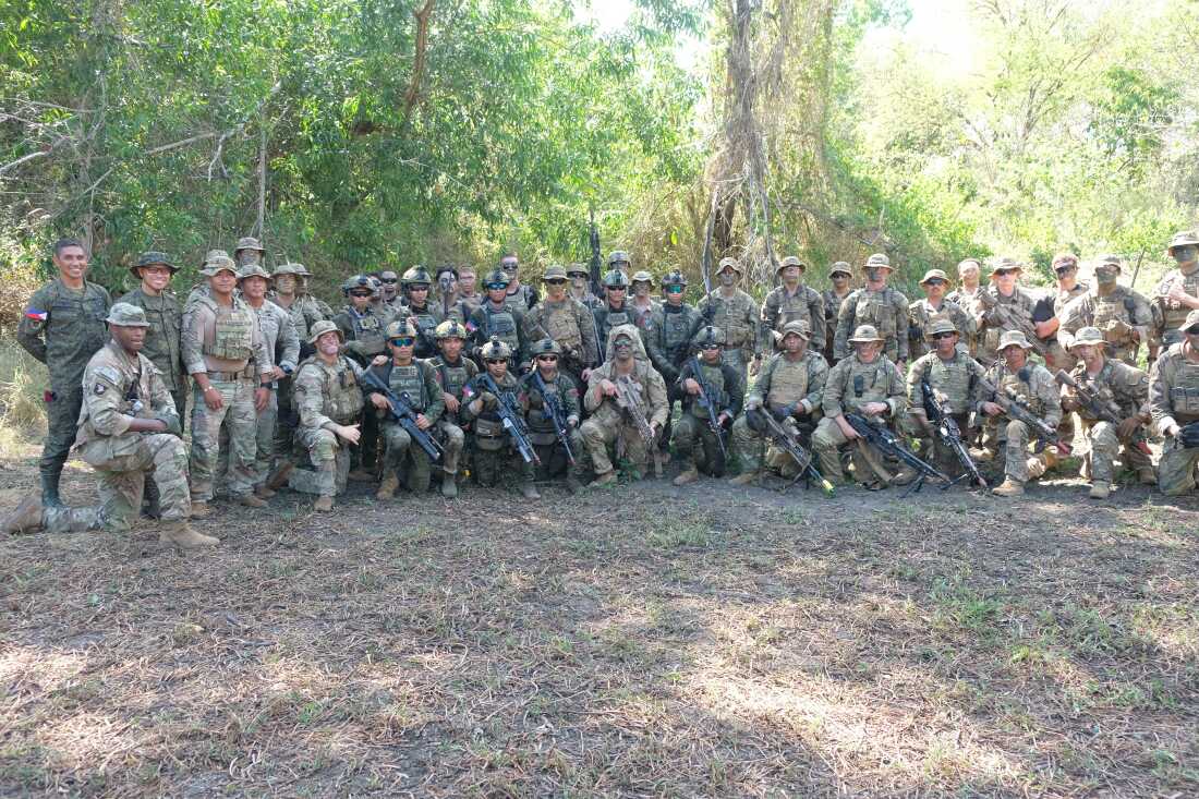 Wearing camouflage-print uniforms, U.S. and Filipino troops stand or kneel in a long row for a photo during annual Balikatan exercises. Some are holding firearms.