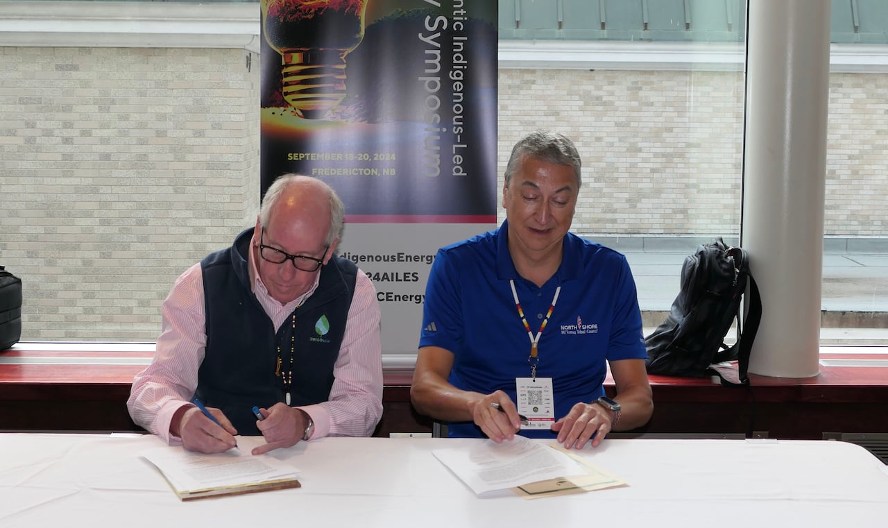Two men sitting together signing documents, with a poster for the Atlantic Indigenous-Led Energy Symposium behind them.