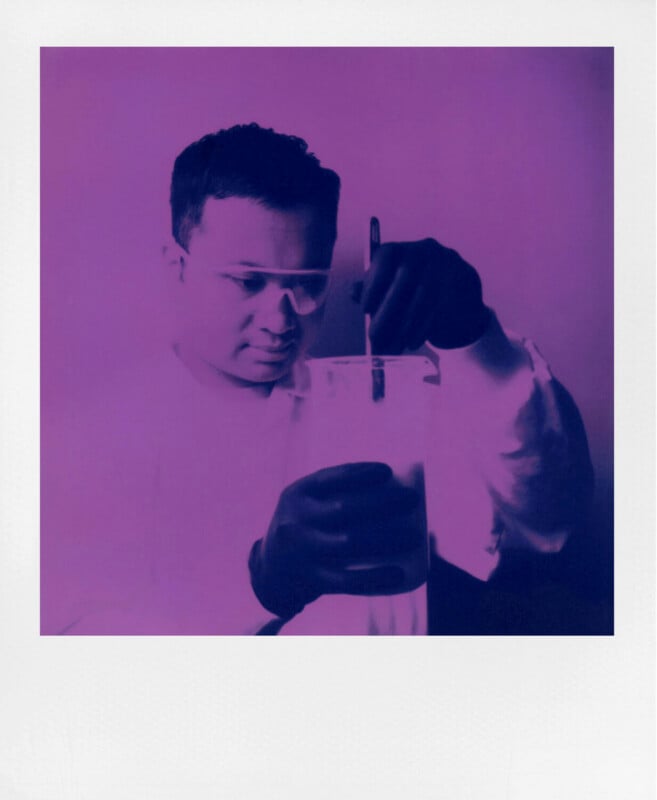 A person wearing safety glasses stirs liquid in a beaker with a glass rod. The image has a strong purple tint and a soft, hazy effect, giving it a futuristic or experimental feel.