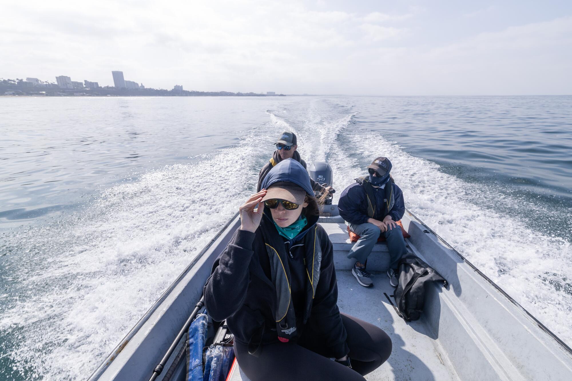 Three people are seen in a motorboat; in the background city buildings are seen along a shoreline.