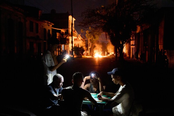 People illuminate themselves with their phones while playing dominoes as a fire set up by residents protesting against prolonged power outages burns on a street in Havana, Cuba, Thursday, May 14, 2026. (AP Photo/Ramon Espinosa)