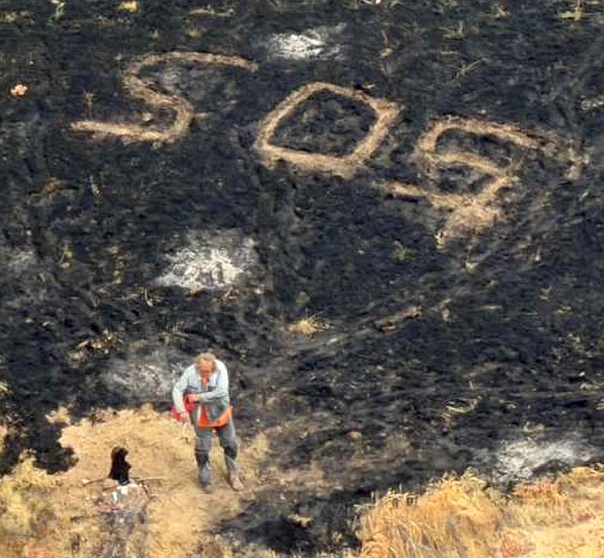 A person stands next to blackened earth with "SOS" carved in.