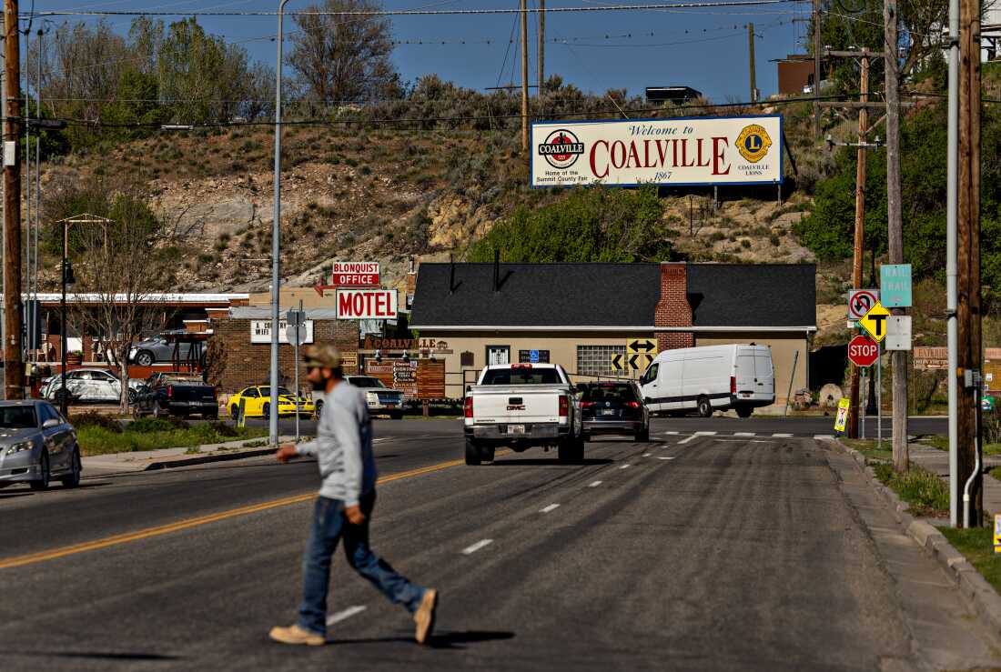 A man crosses a street in a town with a billboard that says "Welcome to Coalville" on the hillside at the end of the street. 
