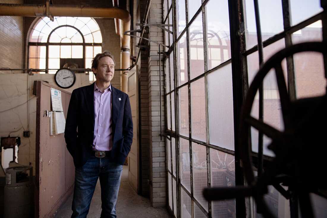 Denver Mayor Mike Johnston stands inside the Cherokee Boiler House, next to a tall, large window made up of a grid of square panes.