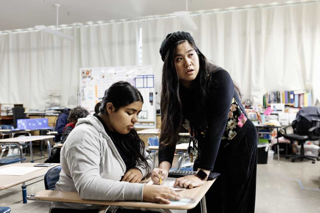 Mary Acebu, a Filipino woman wearing all black with a flowered vest and knitted beret, leans over a desk to help a student at her desk. The student has long brown hair in a ponytail and a gray sweatshirt.