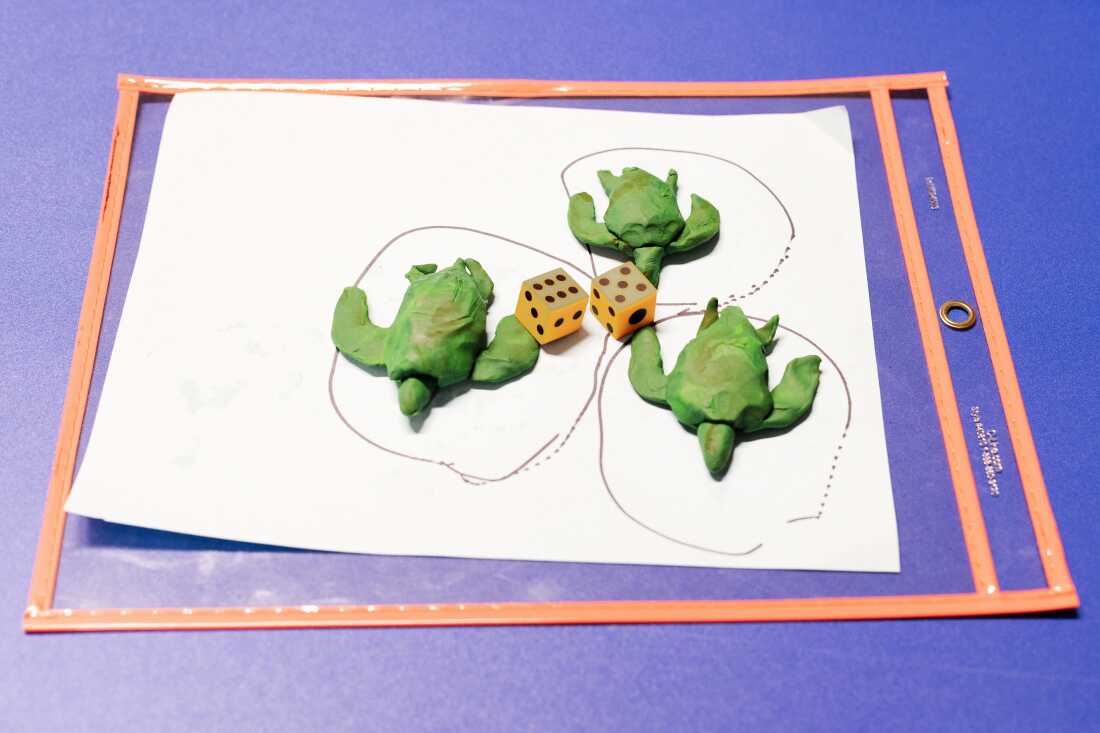 Three green clay turtles sit on a piece of white paper with circles drawn around them. Two dice sit in the middle with the numbers five and six shown.