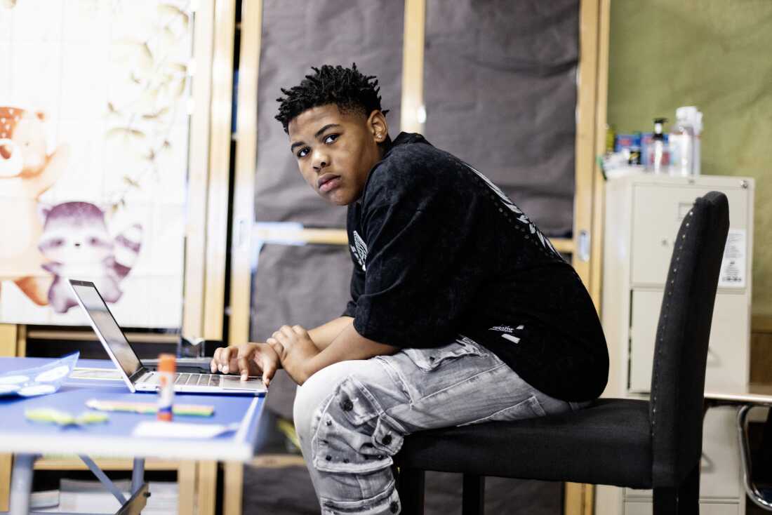 King — a black, eighth grade boy with short locs is wearing a black t-shirt and gray jeans. He looks at the camera while sitting at a laptop working on an assignment in Mary Acebu's class.