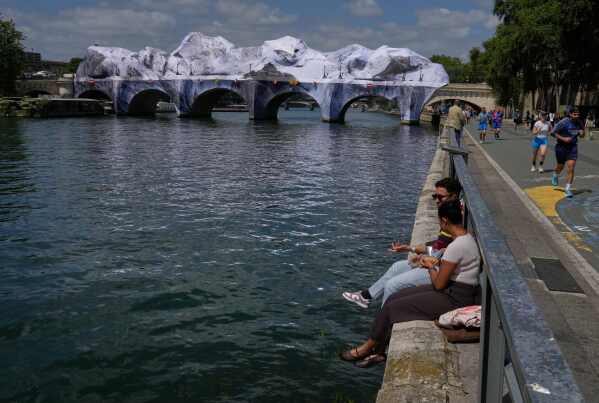 People sit along the Seine river next to "The Pont Neuf Cave," an inflated art installation by French street artist JR, on Paris' oldest bridge, the Pont Neuf, Thursday, May 21, 2026, which will be open to the public from June 6-28. (AP Photo/Michel Euler)