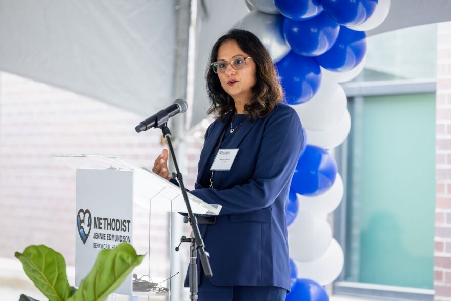 Photo shows the side profile of a brunette woman with glasses in a navy blue suit. Other people near her are out of focus.