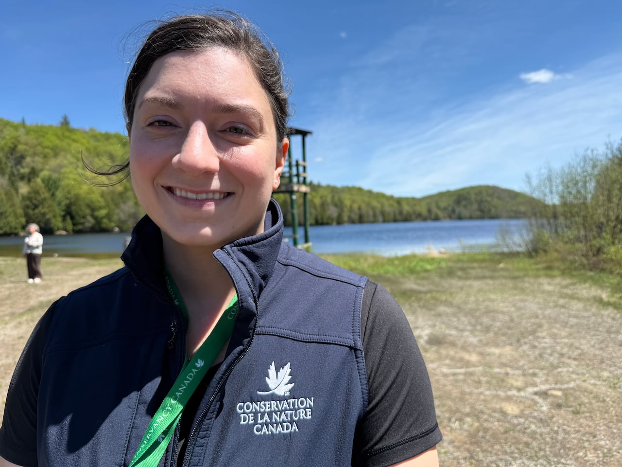 Close up of a woman with dark hair wearing a blue vest with a patch that reads Conservation de la Nature Canada stands on a bleach wit a blue lake behind her