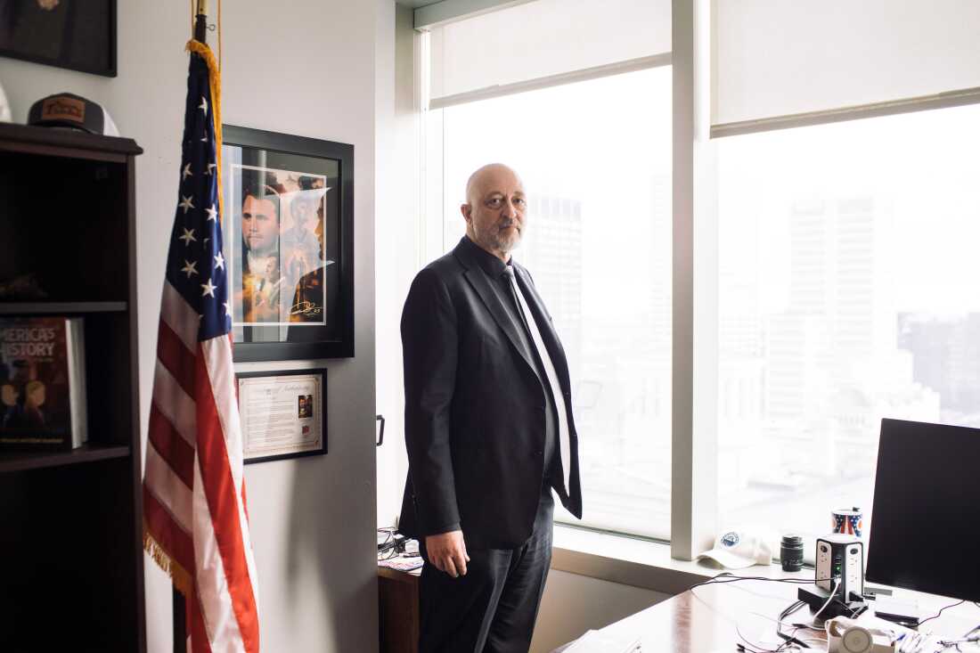 A man in a suit stands in an office next to a U.S. flag.