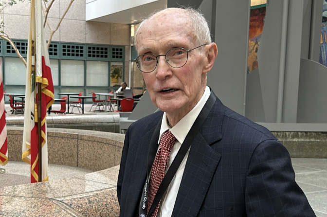 An elderly light skinned man wearing a blue jacket and red spotted tie is sitting in an outside space; an official flag is on a flagpole next to him