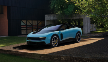 A sleek, modern light blue sports car with black accents is parked on a sunlit concrete platform next to a dark building and surrounded by greenery.